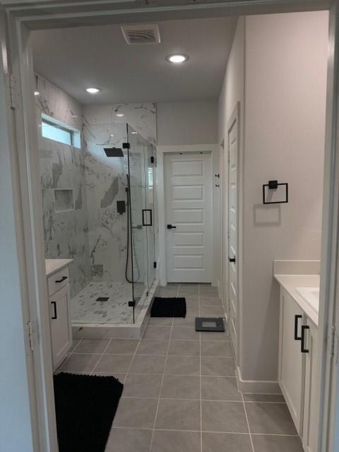 Walk-in shower featuring floor-to-ceiling stone-pattern tile, a glass enclosure, and a wall-mounted shower head, complemented by white cabinetry with matte black hardware and light-toned tile flooring