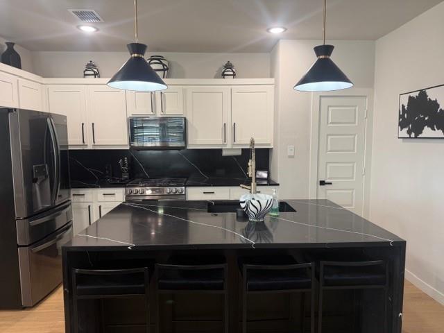 Kitchen featuring white cabinetry, black stone-patterned countertops, a matching central island with built-in sink, stainless steel appliances, and dual pendant lighting