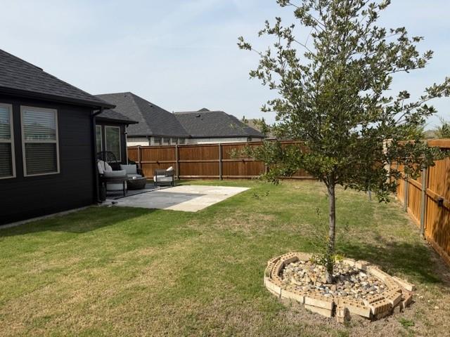 Backyard featuring a concrete patio, wood privacy fencing, grass lawn, and a tree with a decorative stone and brick base