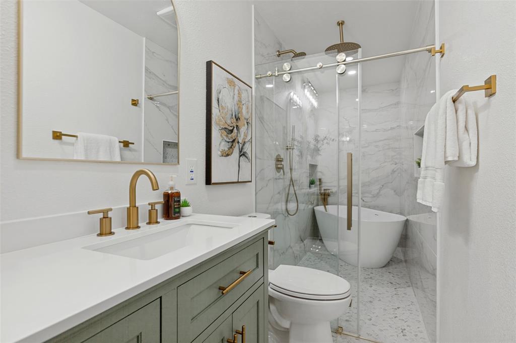 Vanity with solid surface countertop, green cabinetry, brass hardware, arched wall mirror, and a walk-in wet room featuring stone-finish wall tile, a freestanding soaking tub, and a rainfall showerhead behind a sliding glass door
