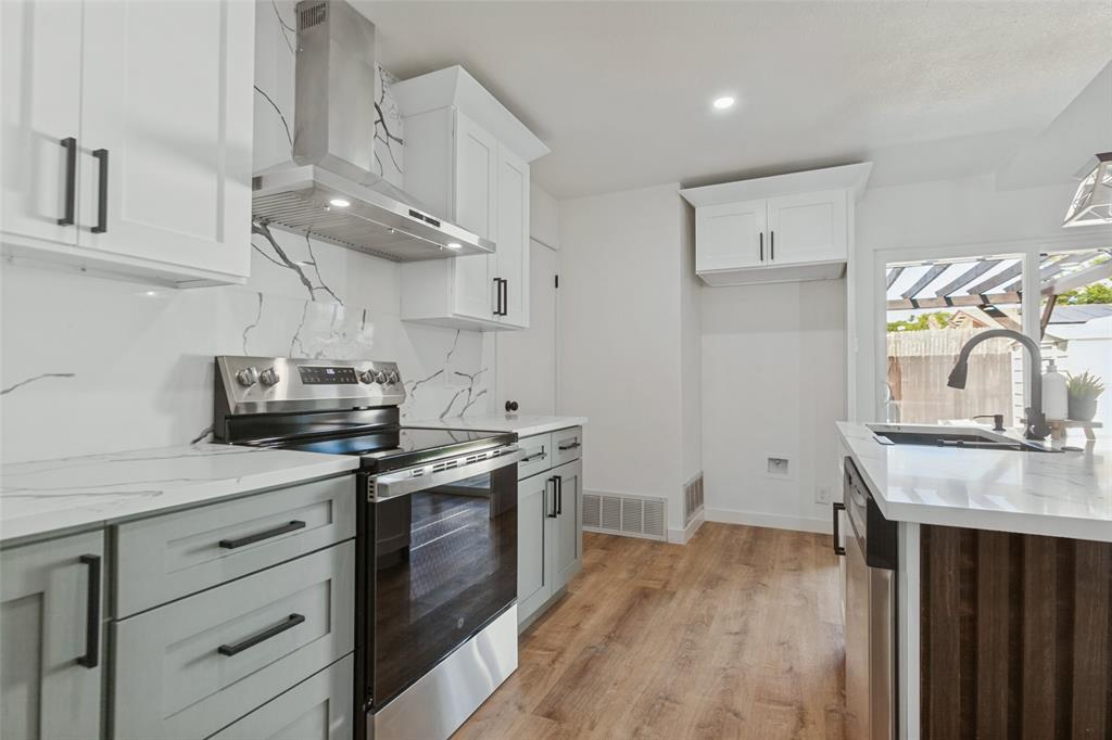 Kitchen featuring white upper cabinetry, soft grey lower cabinetry, stone-patterned backsplash, stainless steel chimney-style range hood, and wood-finish flooring