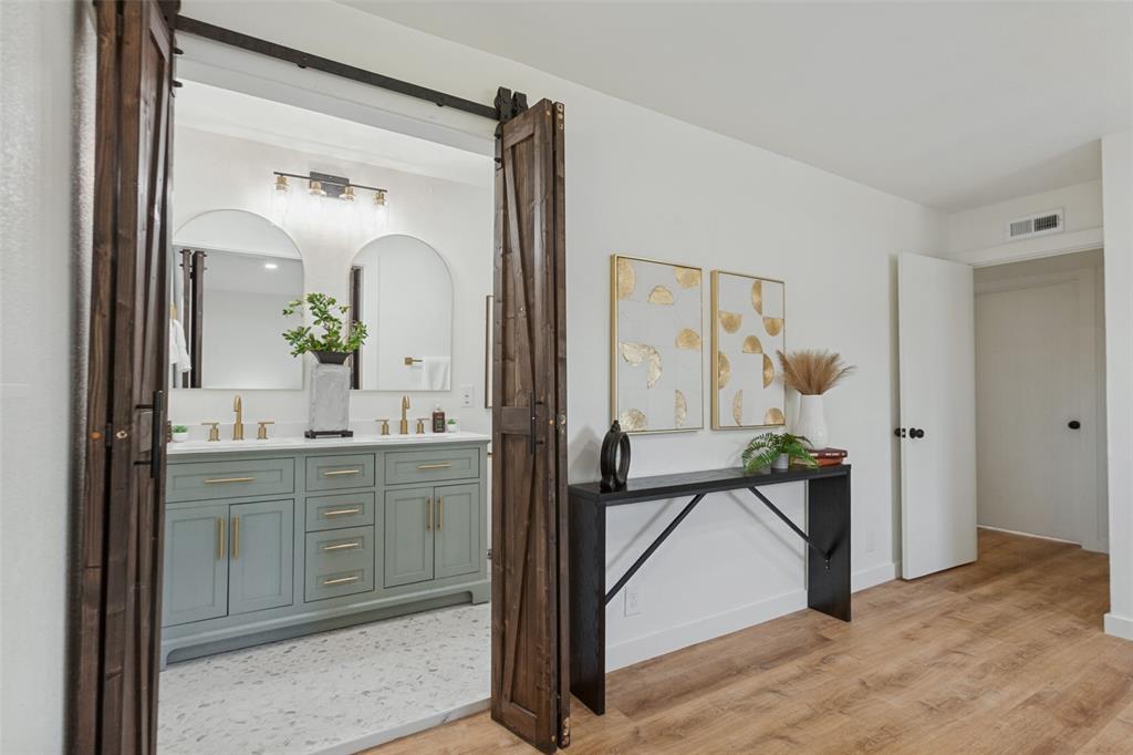 Double vanity with a light-toned cabinet, brass hardware, arched mirrors, and wood-finish flooring, featuring a dark-stained sliding barn door entry