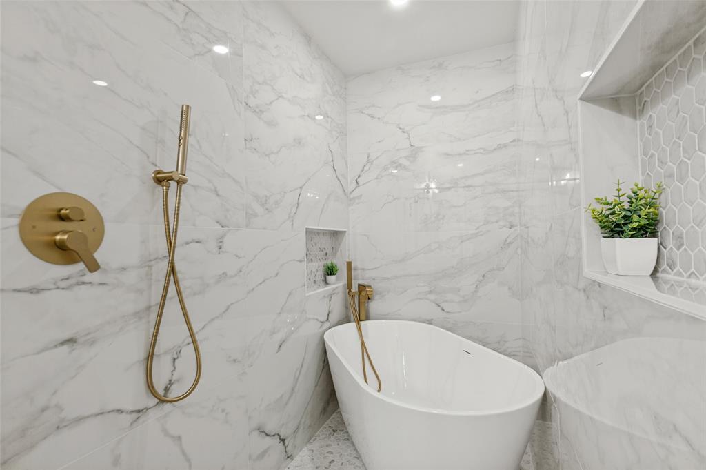 Freestanding soaking tub featuring a metallic handheld faucet, wall-integrated shelving, and floor-to-ceiling large-format stone-finish tile surround