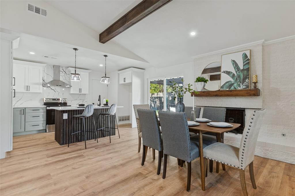 Open-concept interior featuring vaulted ceilings with exposed wood beams, wood-finish flooring, kitchen island with vertical slat detailing, and a fireplace with a textured surround and heavy timber mantel