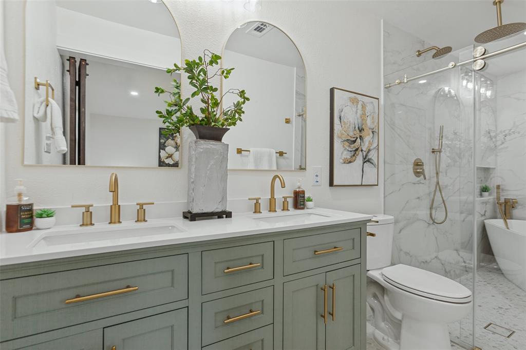 Double vanity with green cabinetry and quartz countertop, dual arched wall mirrors, brass-finish fixtures, walk-in glass-enclosed shower, and freestanding soaking tub