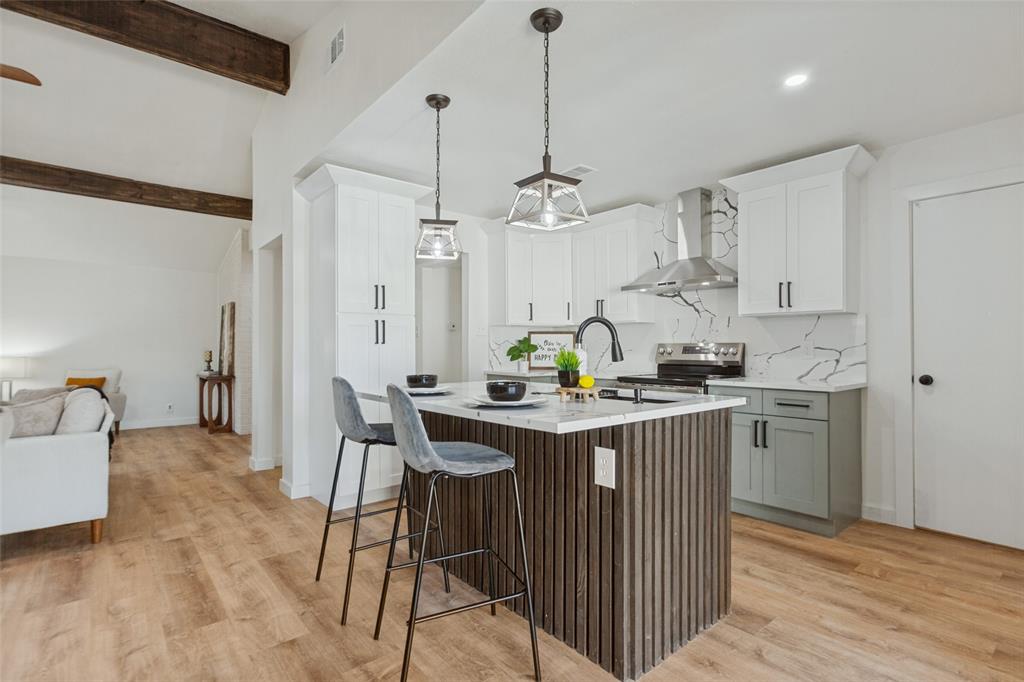 Kitchen featuring wood-finish flooring, a central island with vertical wood slat detailing, stone-patterned backsplash, white shaker cabinetry, and stainless steel range hood