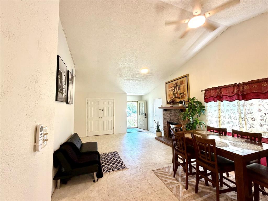 Living and dining area featuring vaulted ceilings, a ceiling fan with integrated lighting, a brick fireplace with a wood mantle, and light-toned tile flooring Living and dining area featuring vaulted ceilings, a ceiling fan with integrated lighting, a brick fireplace with a wood mantle, and light-toned tile flooring
