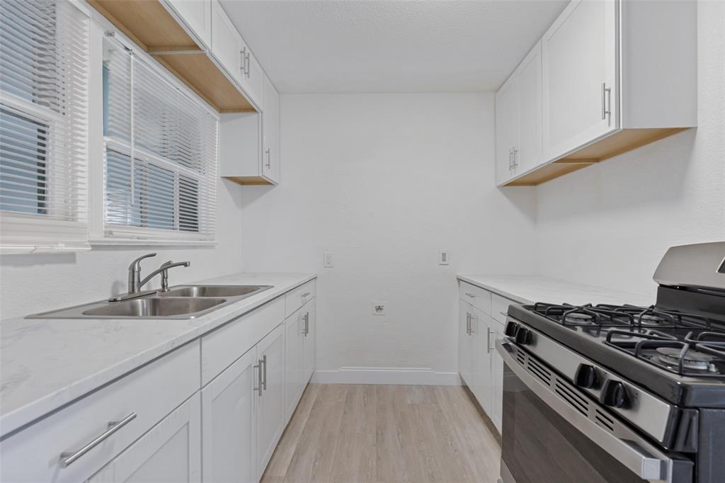 Kitchen featuring white cabinetry, light-toned stone-finish countertops, wood-finish flooring, stainless steel double-basin sink, and a built-in gas range