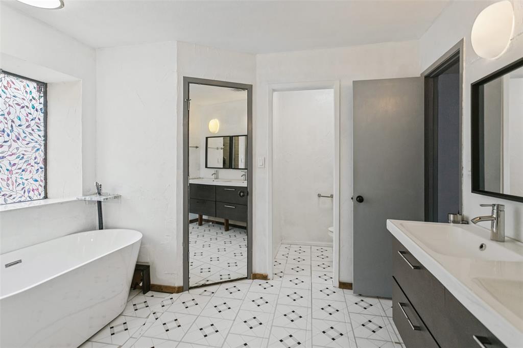 Freestanding soaking tub, dual-sink vanity with dark cabinetry, patterned tile flooring, and decorative leaded glass window