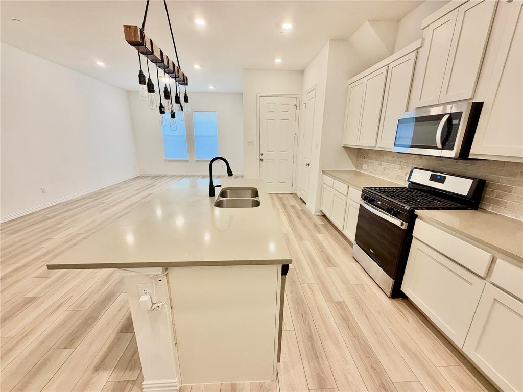 Kitchen featuring wood-finish flooring, white shaker-style cabinetry, a central island with a double-basin sink, quartz-style countertops, and a linear pendant light fixture