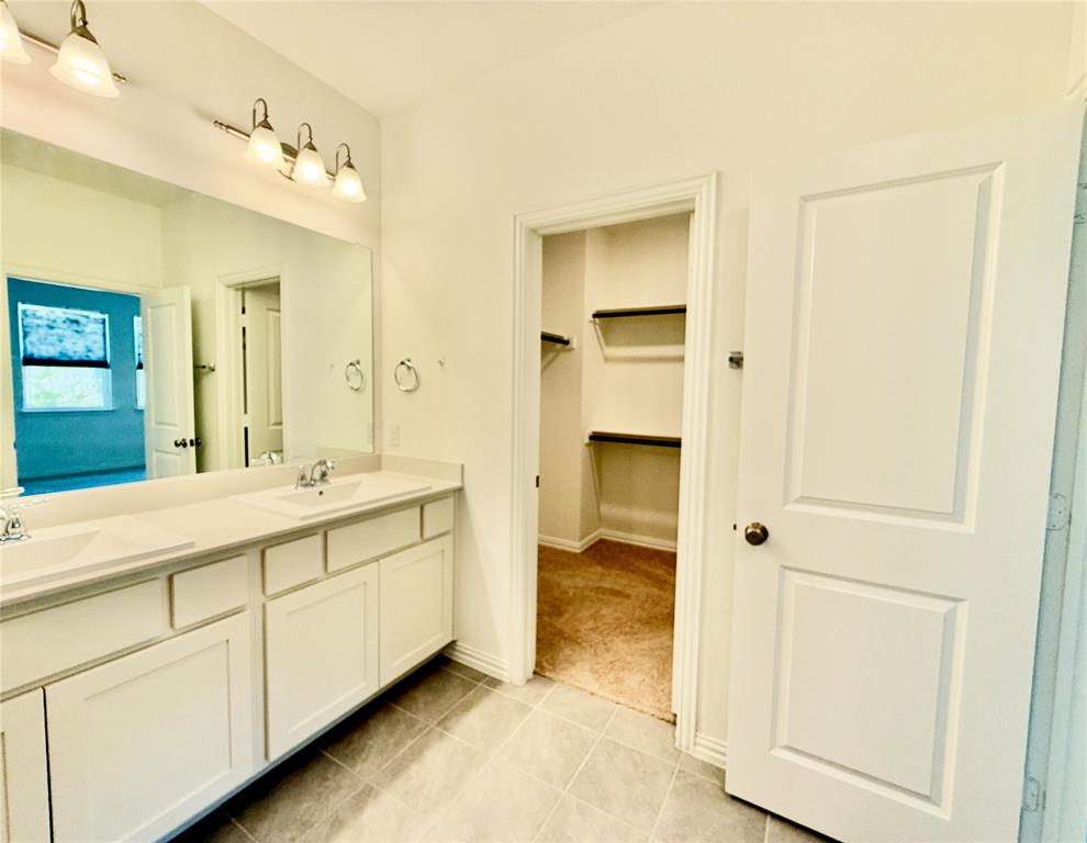 Dual-sink vanity with white cabinetry, integrated countertop, and oversized wall-mounted mirror, accented by dual three-light fixtures and tile flooring