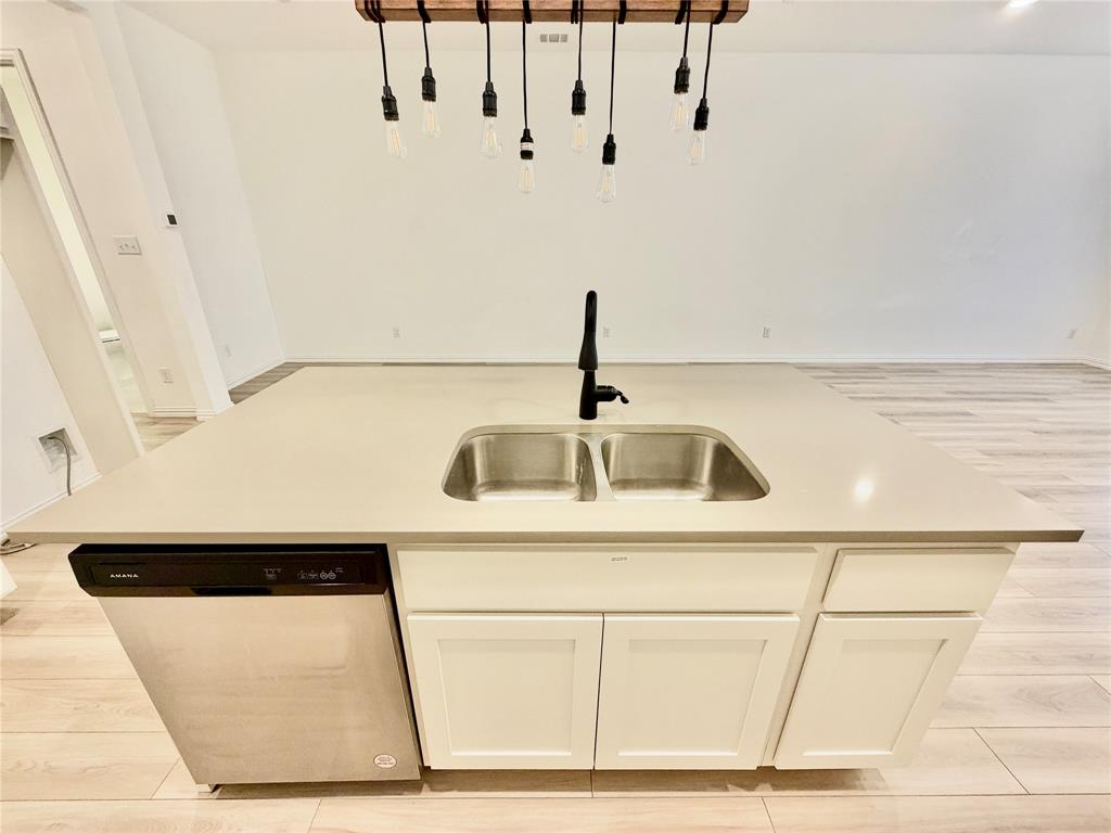 Kitchen island with light-toned solid surface countertop, stainless steel undermount double-basin sink, matte black gooseneck faucet, integrated stainless steel dishwasher, and suspended wood-beam pendant lighting with exposed filament bulbs over wood-finish flooring