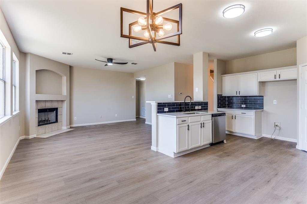 Open-concept interior featuring wood-finish flooring, a kitchen island with a dark tile backsplash, white cabinetry, a geometric statement chandelier, and a recessed fireplace with a neutral tile surround