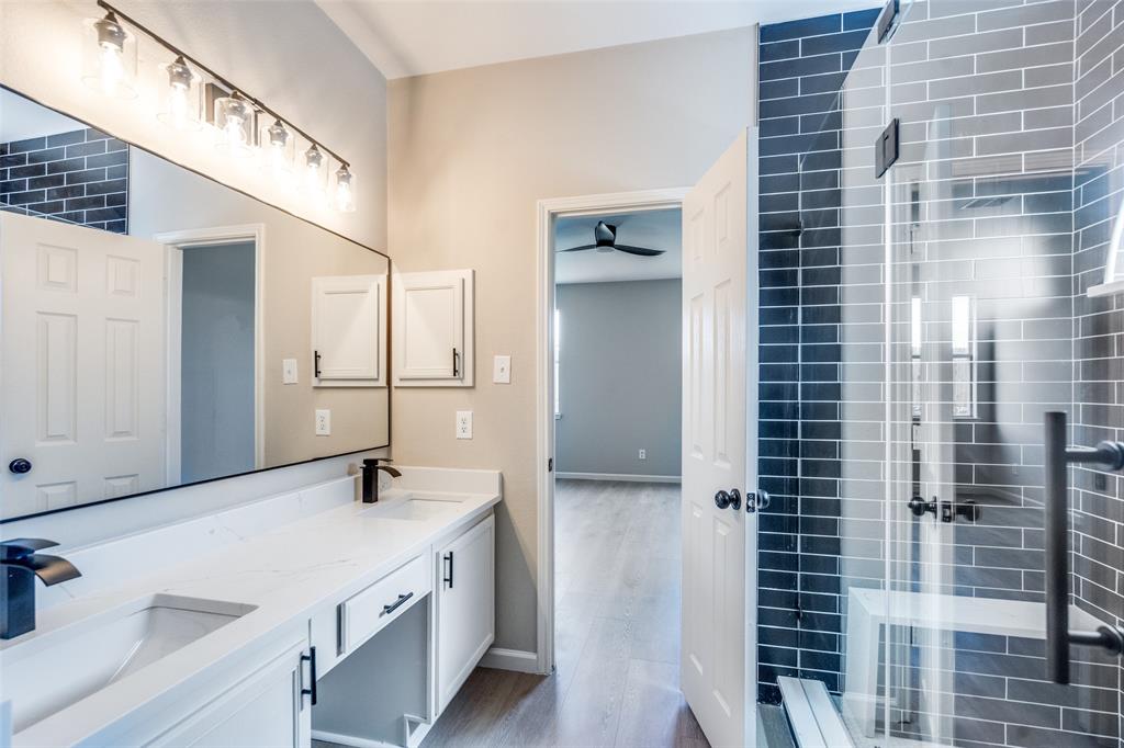 Dual-sink vanity with quartz countertop, walk-in shower featuring glass enclosure and dark ceramic wall tile, full-width vanity mirror, and matte black hardware