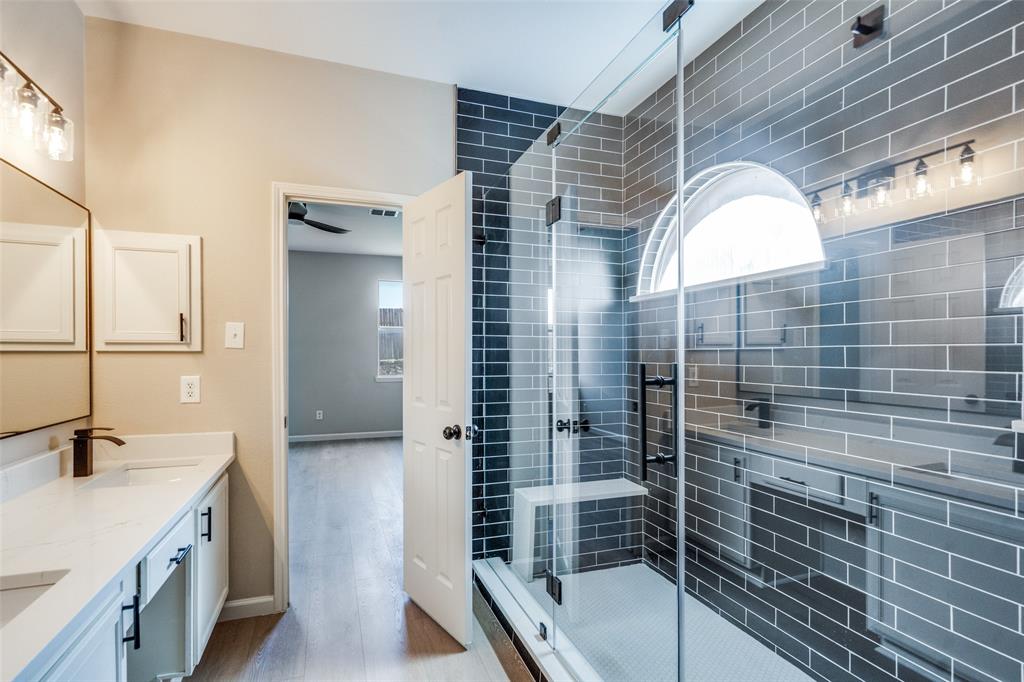 Double vanity with stone-style countertops, glass-enclosed shower featuring subway tile surround, built-in shower bench, and arched transom window