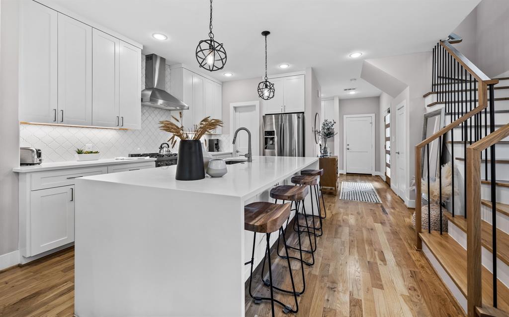 Kitchen featuring wall chimney range hood, light hardwood / wood-style flooring, hanging light fixtures, an island with sink, and stainless steel fridge Kitchen featuring wall chimney range hood, light hardwood / wood-style flooring, hanging light fixtures, an island with sink, and stainless steel fridge