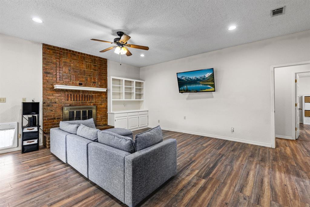 Living area featuring wood-finish flooring, floor-to-ceiling brick fireplace with wood mantel, built-in shelving, and ceiling fan with integrated lighting