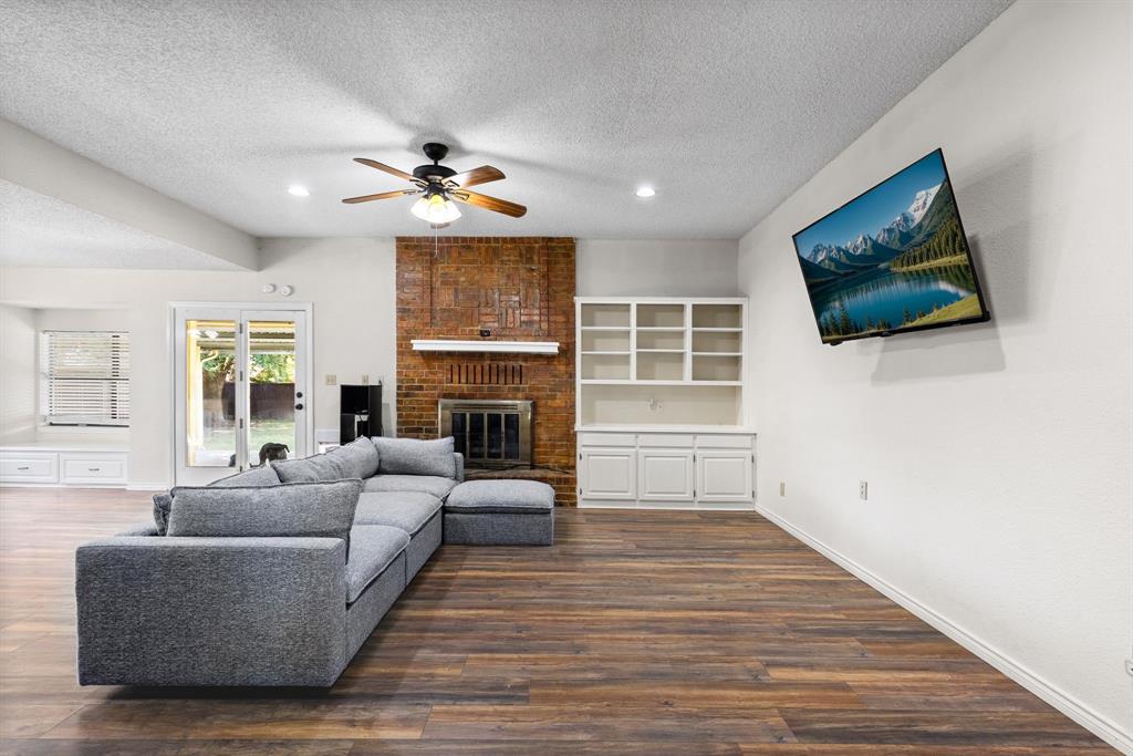 Living area featuring wood-finish flooring, floor-to-ceiling brick fireplace with wood mantel, built-in white shelving, recessed lighting, and sliding glass doors