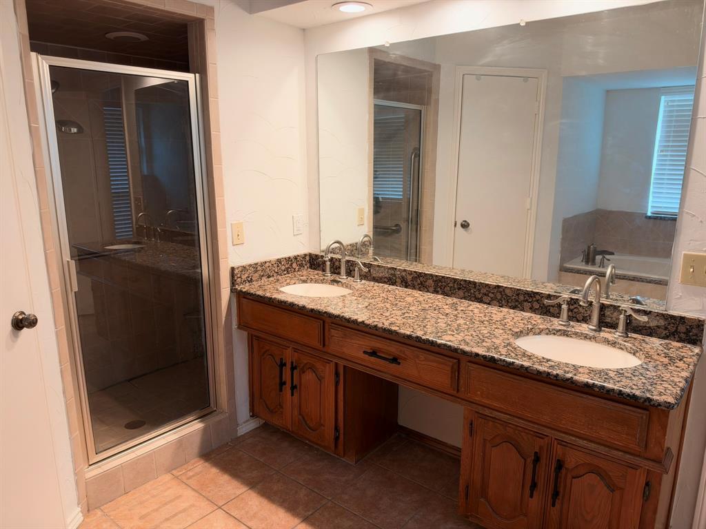 Dual-sink vanity featuring stone-finish countertops, wood cabinetry, inset oval basins, and an expansive wall-to-wall mirror, complemented by a walk-in shower enclosure with a glass door and tiled flooring