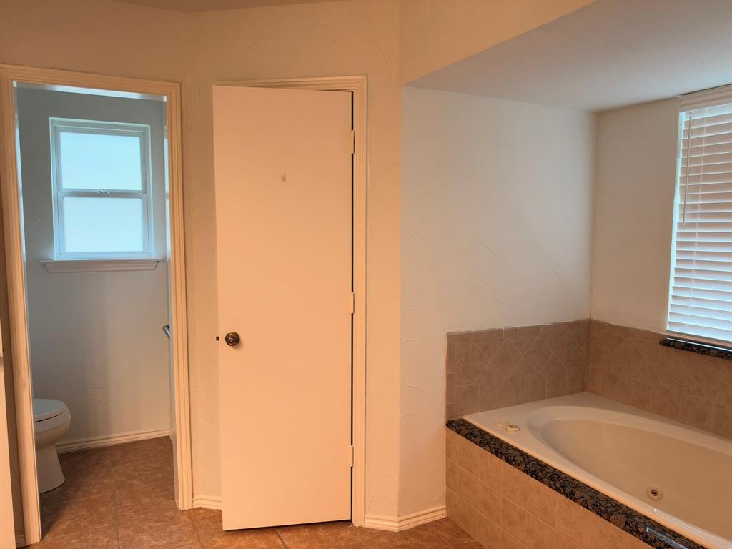 Jetted soaking tub with tile surround, stone-finish ledge, and blinds, featuring tile flooring and an enclosed water closet
