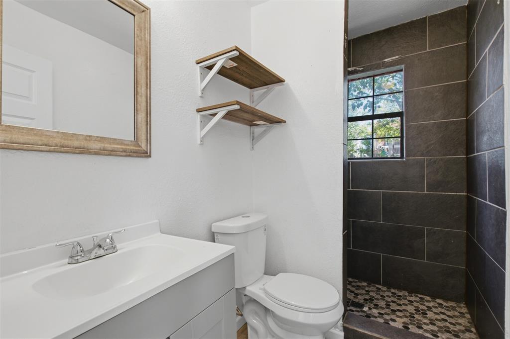 Bathroom vanity with a solid-surface countertop and undermount sink, wall-mounted open shelving, framed mirror, and a walk-in shower with large-format rectangular wall tile and mosaic floor tile