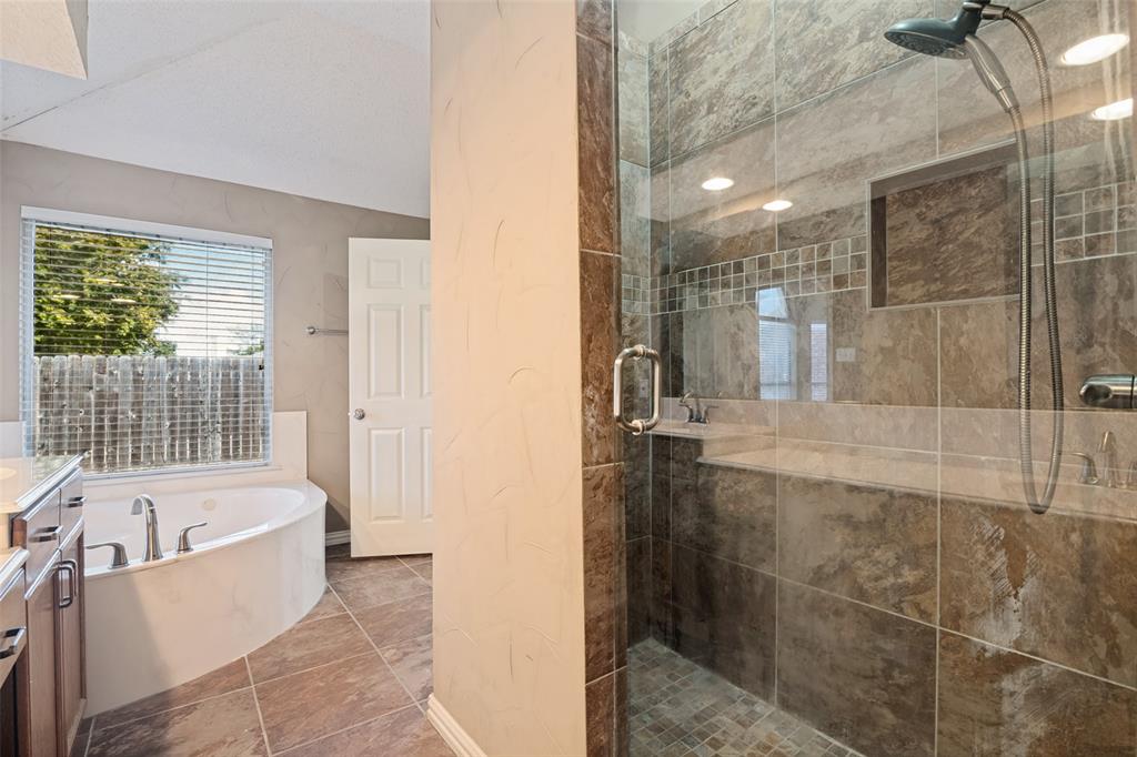 Walk-in shower with stone-tile surround, glass enclosure, and built-in recessed niche; deep soaking jetted tub; tile flooring; and wood-finish cabinetry