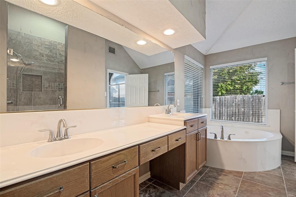 Dual-sink vanity with wood cabinetry and light-toned solid surface countertops, glass-enclosed walk-in shower with tiled surround, and jetted soaking tub