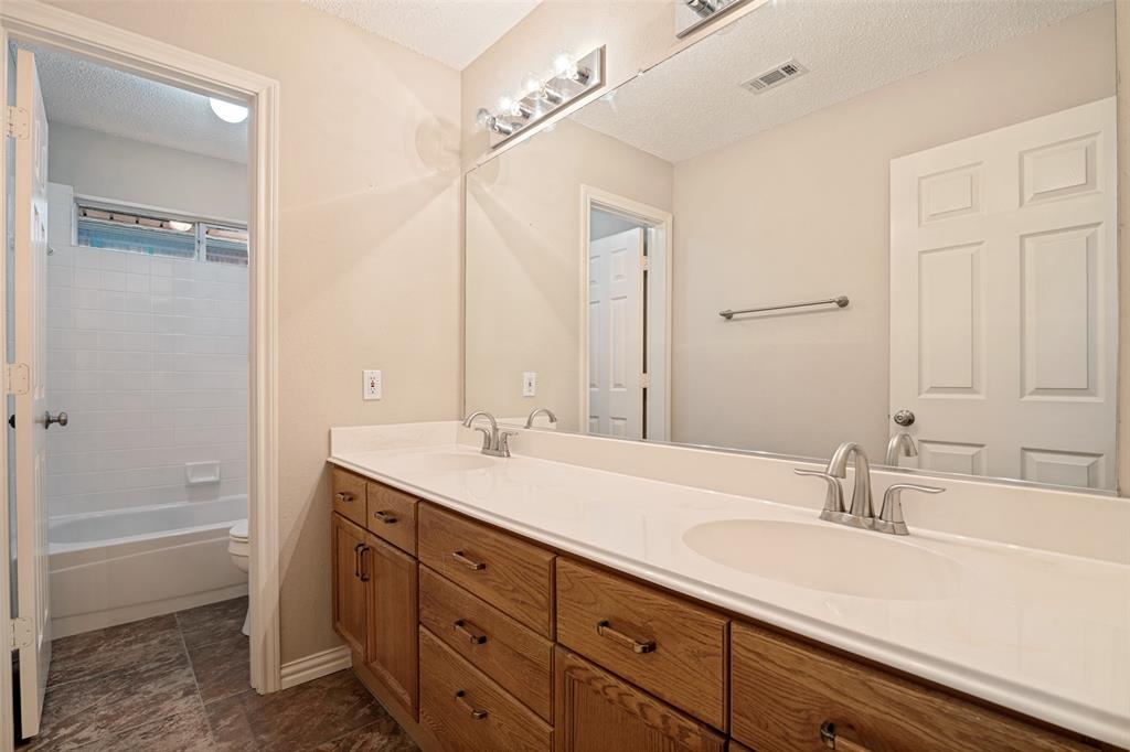 Dual-sink vanity with wood-finish cabinetry, solid surface countertop, large wall-mounted mirror, and brushed-finish faucets, featuring a separate water closet area with a tub-shower combination and tiled flooring