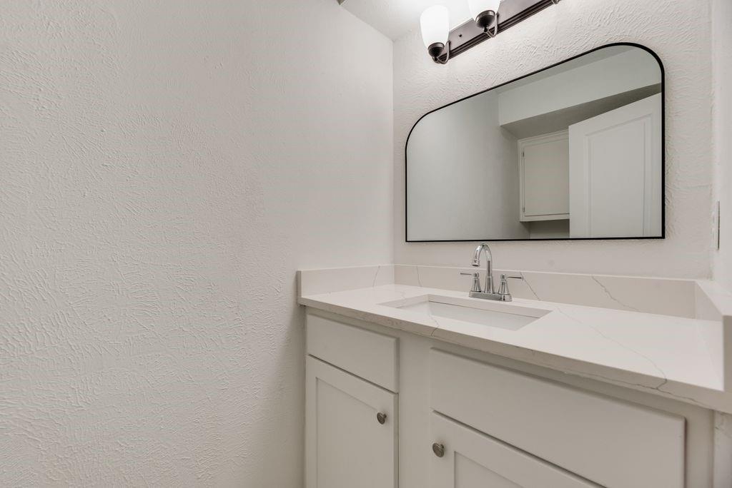 Vanity featuring white cabinetry, quartz countertop, integrated rectangular sink, chrome hardware, and an arched-frame wall mirror