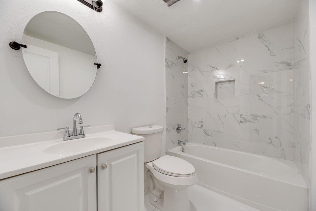 Bathroom featuring a white vanity cabinet with an integrated sink, wall-mounted oval mirror, and a shower-tub combination with white tile surrounds and a built-in recessed niche Bathroom featuring a white vanity cabinet with an integrated sink, wall-mounted oval mirror, and a shower-tub combination with white tile surrounds and a built-in recessed niche
