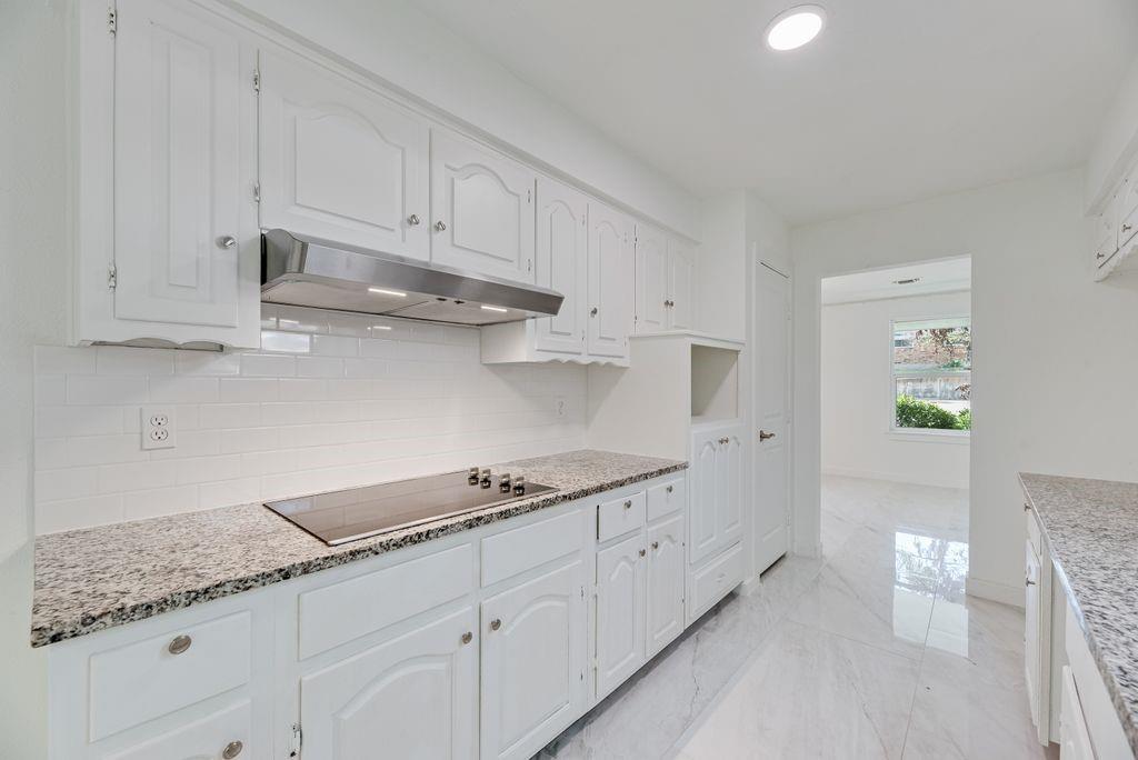 Kitchen featuring white cabinetry, stone-finish countertops, subway tile backsplash, stainless steel range hood, and large-format tile flooring Kitchen featuring white cabinetry, stone-finish countertops, subway tile backsplash, stainless steel range hood, and large-format tile flooring