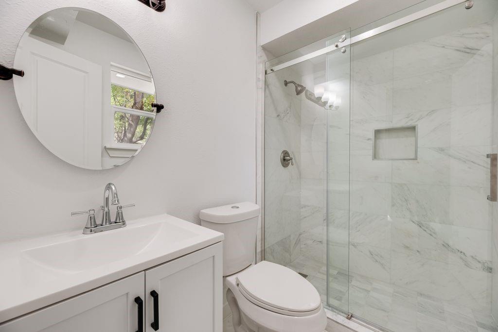Bathroom featuring a white vanity cabinet, solid-surface countertop, round wall-mounted mirror, and a walk-in shower with stone-style tile surround and a sliding glass door