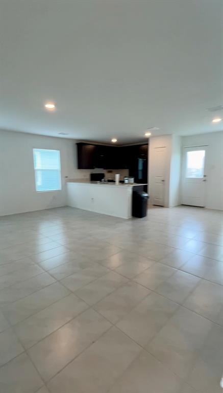 Open-concept interior featuring large-format tile flooring, recessed ceiling lighting, kitchen with dark cabinetry, stone-surface breakfast bar, and panel-style entry door