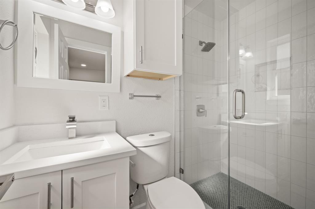 White vanity with a rectangular sink, wall-mounted mirror, white cabinetry, a glass-enclosed shower with tiled surround, and mosaic tile shower flooring