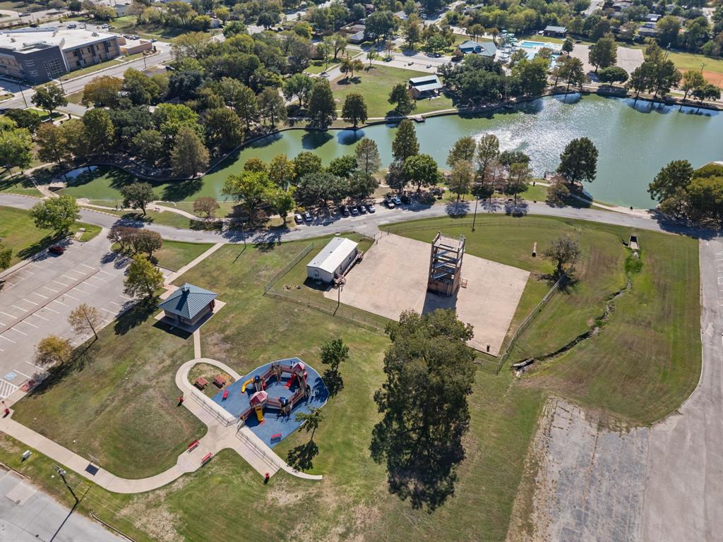 Aerial view of community park grounds featuring a concrete-surfaced area, a dedicated playground structure with cushioned surfacing, a multi-vehicle parking lot, and bordering natural water features Aerial view of community park grounds featuring a concrete-surfaced area, a dedicated playground structure with cushioned surfacing, a multi-vehicle parking lot, and bordering natural water features