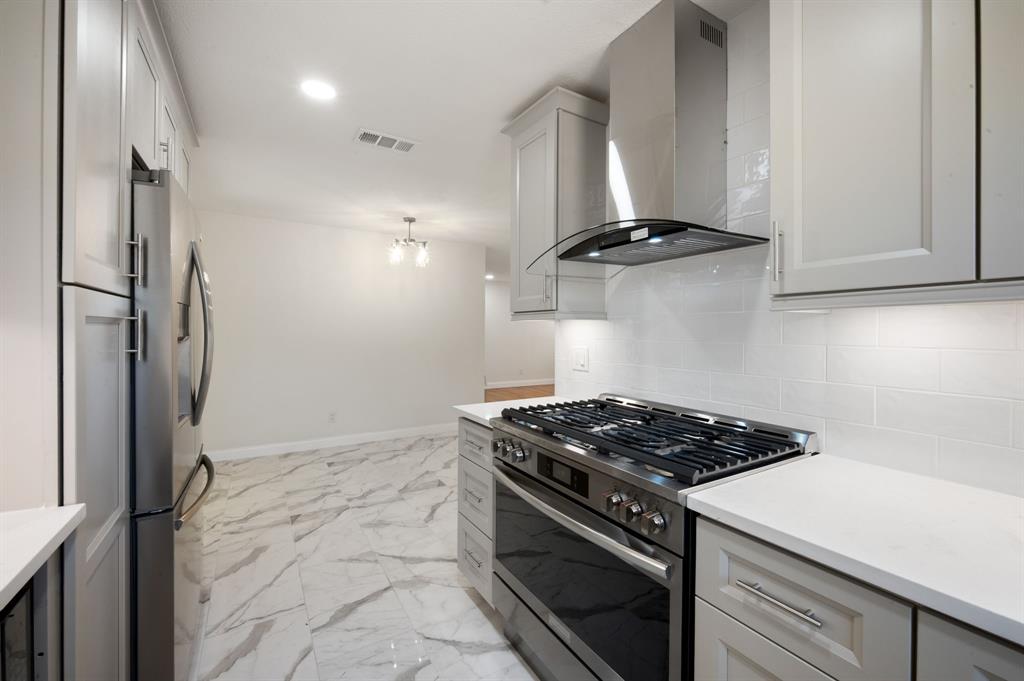 Kitchen featuring light-toned shaker cabinetry, stainless steel gas range with overhead vent hood, white subway tile backsplash, solid surface countertops, and stone-finish flooring