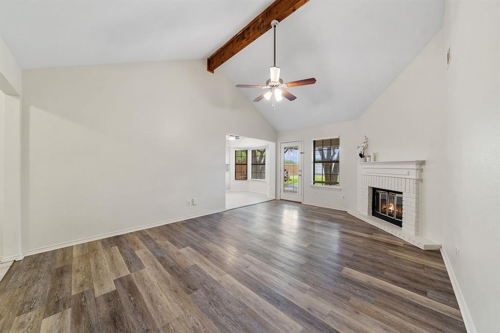 Vaulted ceiling featuring a prominent wooden support beam, LVP flooring, white brick fireplace with a mantel, and a ceiling fan with integrated lighting Vaulted ceiling featuring a prominent wooden support beam, LVP flooring, white brick fireplace with a mantel, and a ceiling fan with integrated lighting