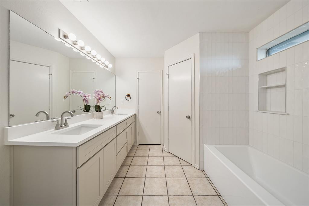 Bathroom featuring an expansive vanity with dual undermount sinks, solid-surface countertop, full-width mirror, tile flooring, and a tub-shower combination with built-in wall shelving