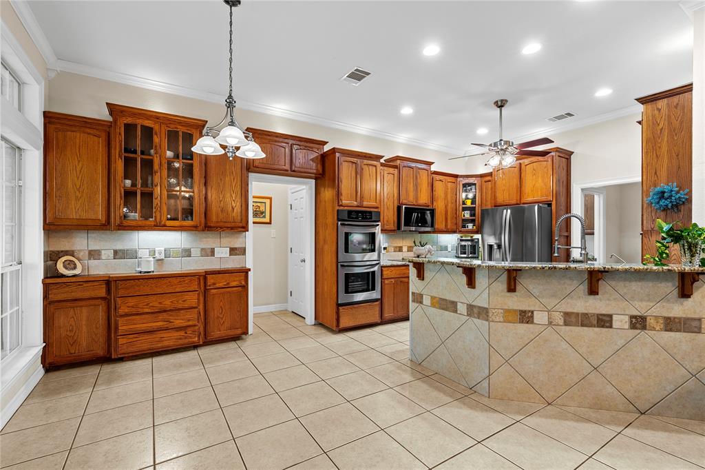 Kitchen featuring stained wood cabinetry, stone-tiled backsplash, built-in double wall oven, peninsula with stone-tiled base, and ceramic tile flooring Kitchen featuring stained wood cabinetry, stone-tiled backsplash, built-in double wall oven, peninsula with stone-tiled base, and ceramic tile flooring