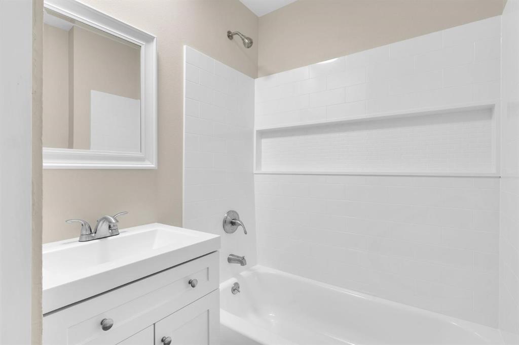 White vanity with a rectangular sink, brushed nickel dual-handle faucet, framed wall mirror, and tub-shower combination featuring white tile surround with a built-in recessed shelf