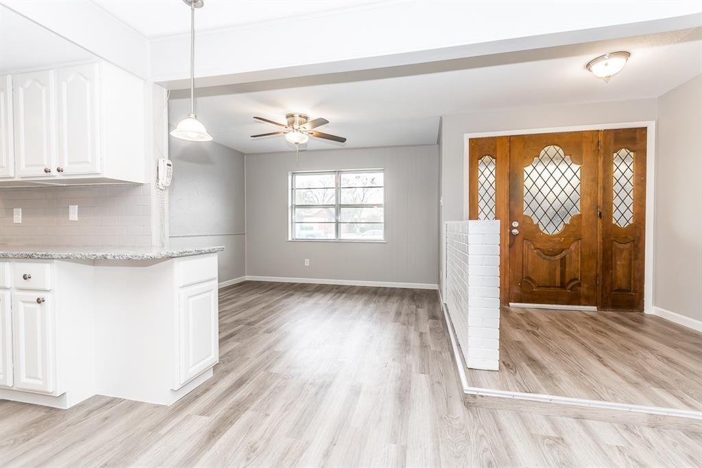 Solid wood entry door with decorative lattice glass inlays, wood-finish flooring, white cabinetry with stone-finish countertops, ceiling fan with light kit, and white painted brick divider wall