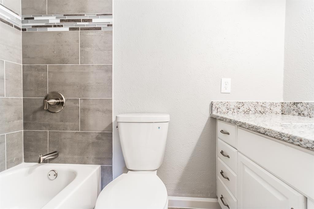 Bathroom featuring a vanity with a light-toned stone countertop, white cabinetry with silver-tone hardware, a bathtub with grey tile surround, and a decorative glass and stone mosaic accent border