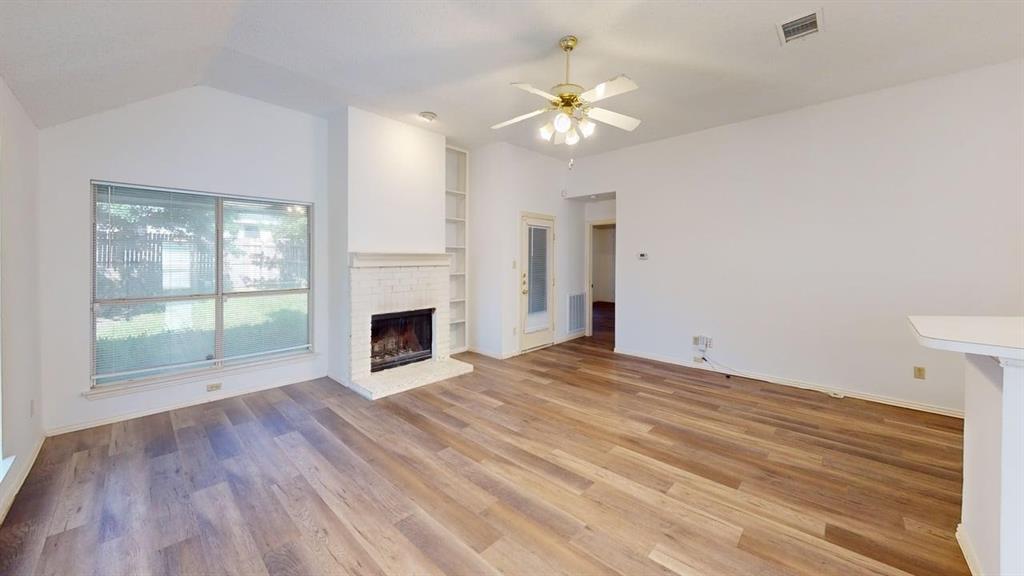 Living area featuring vaulted ceilings, wood-finish flooring, a painted brick fireplace, built-in shelving, and a ceiling fan with integrated lighting