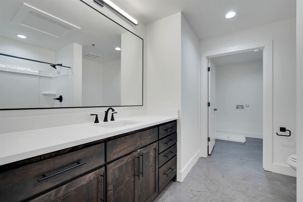 Bathroom featuring custom dark-wood cabinetry, white solid-surface countertop, matte black fixtures, full-width mirror with integrated lighting, and polished concrete flooring
