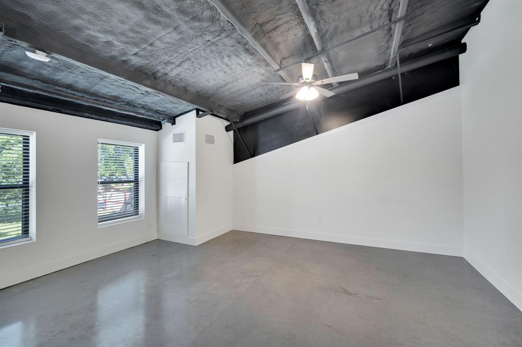 Industrial-style space featuring exposed painted ceiling structure, concrete flooring, dual-pane windows, and a ceiling-mounted light fixture with fan