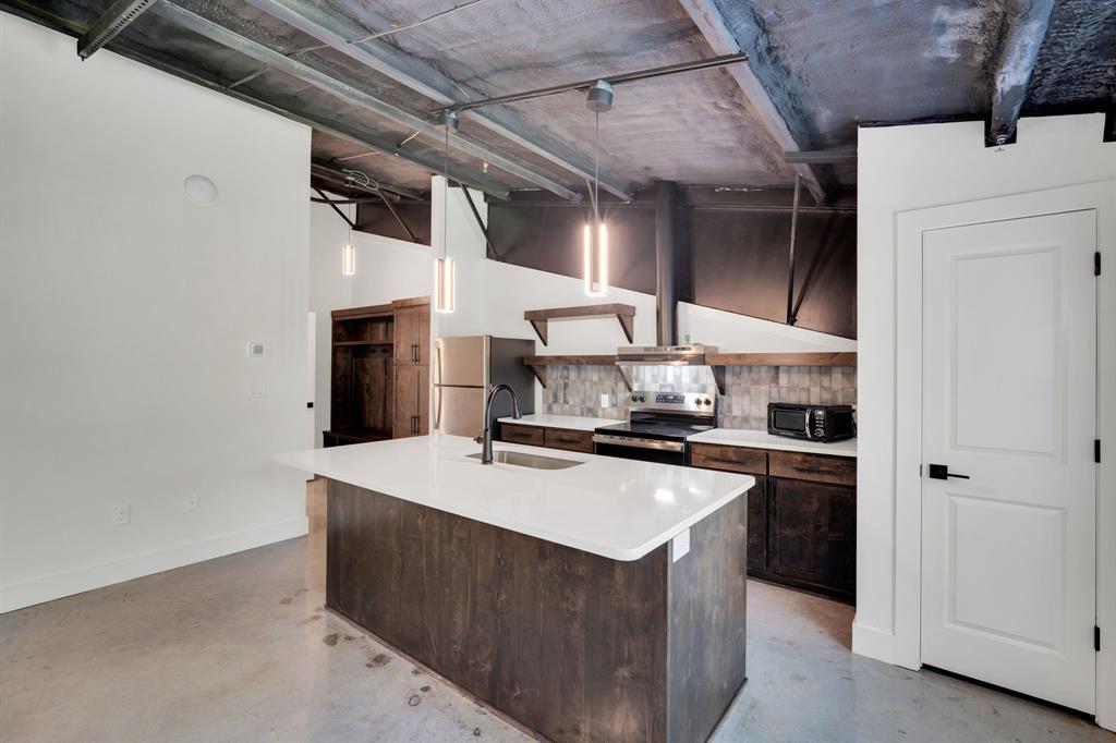 Industrial-style kitchen featuring an exposed concrete ceiling, dark wood-finish cabinetry, quartz countertops, a central island with an integrated sink, and cylinder pendant lighting