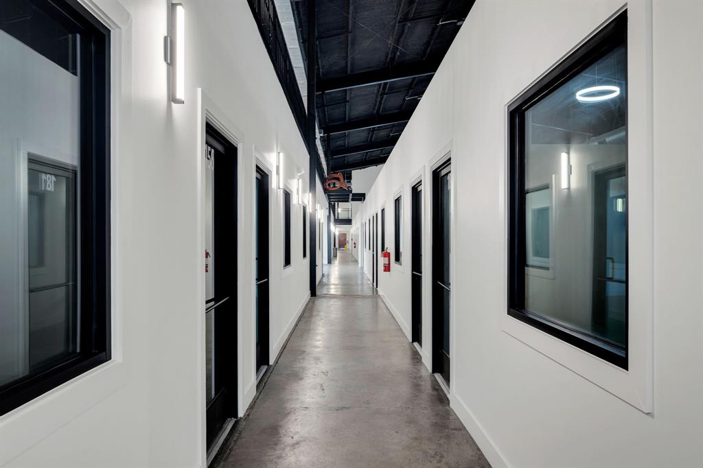 Industrial-style corridor featuring polished concrete flooring, exposed high-ceiling trusses, crisp white walls, floor-to-ceiling glass-paneled doors, and modern wall-mounted linear light fixtures