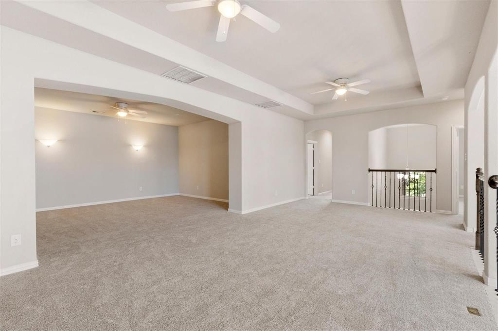 Expansive room featuring recessed tray ceilings, neutral carpeting, three ceiling fans, arched entryways, and decorative metal balusters