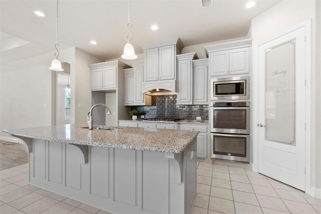 Kitchen featuring light gray cabinetry, expansive stone-topped island, stainless steel double wall ovens, glass-tiled backsplash, and pantry door with frosted glass panel