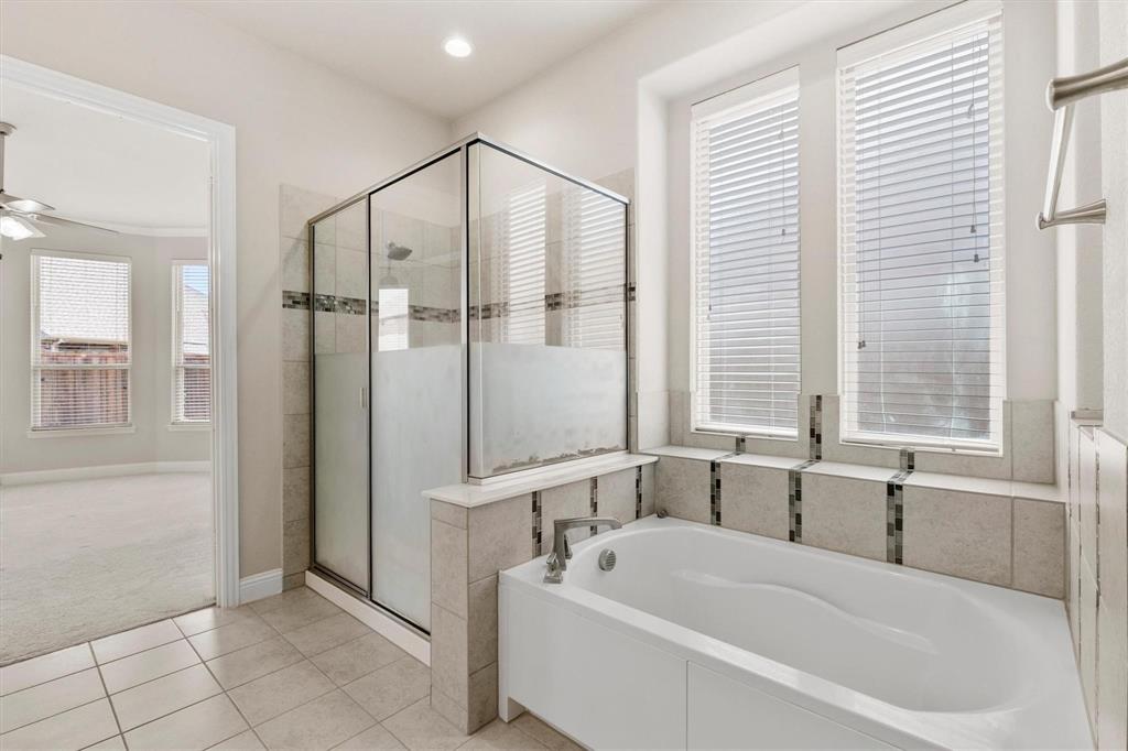 Bathroom featuring a glass-enclosed shower, a deep soaking tub with tile surround, neutral ceramic tile flooring, and dual windows with integrated blinds
